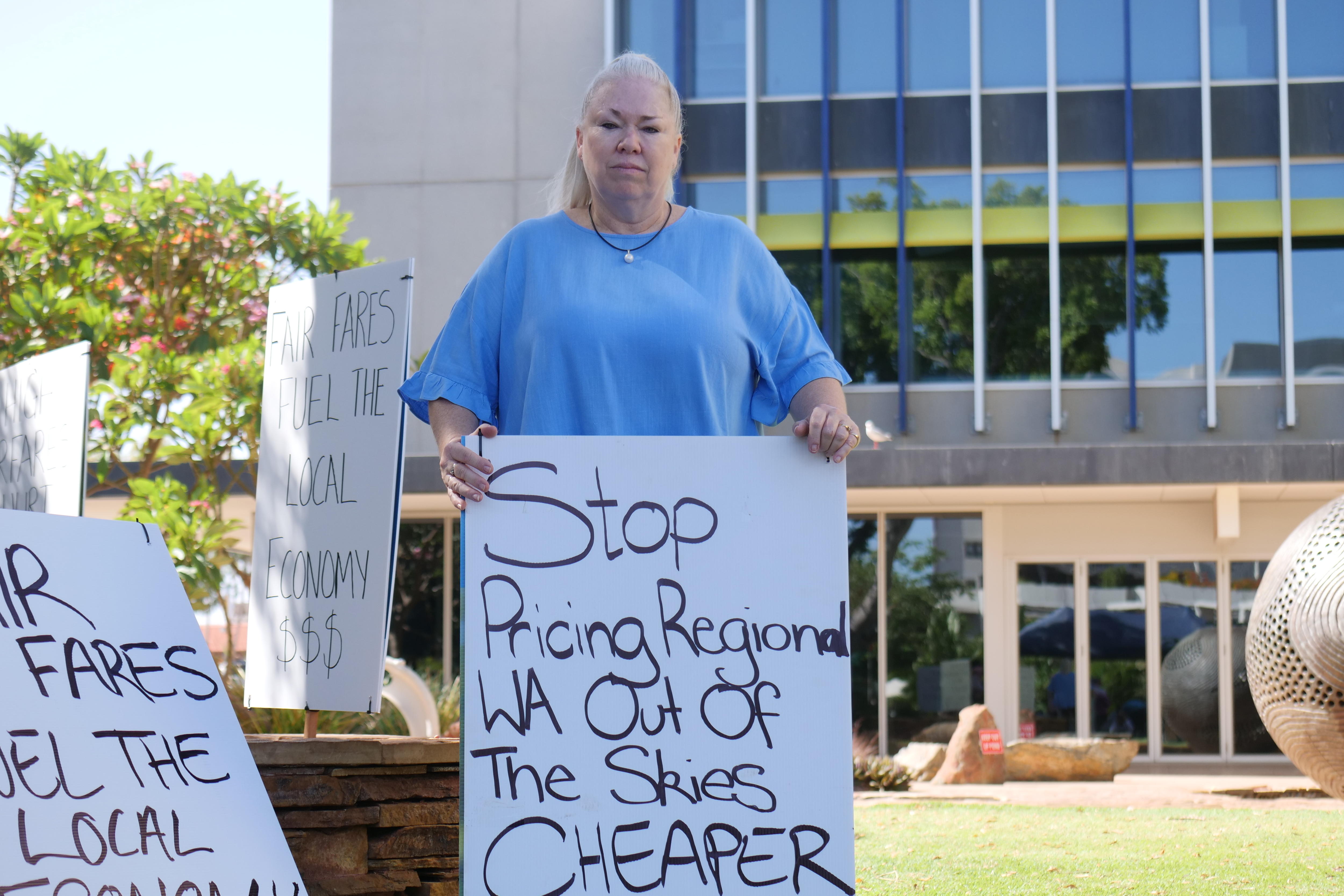 Una mujer rubia de mediana edad sostiene un cartel que promueve tarifas aéreas más baratas mientras se encuentra en una zona de césped cerca de un gran edificio.