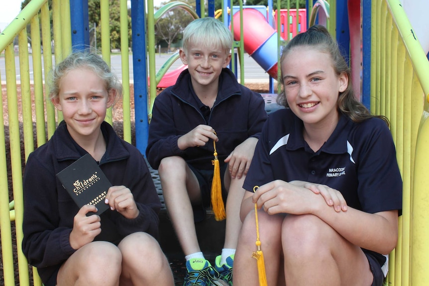 Three children sit next to each other on playground stairs. Two are holding the tassels from their caps, one a learning diary.