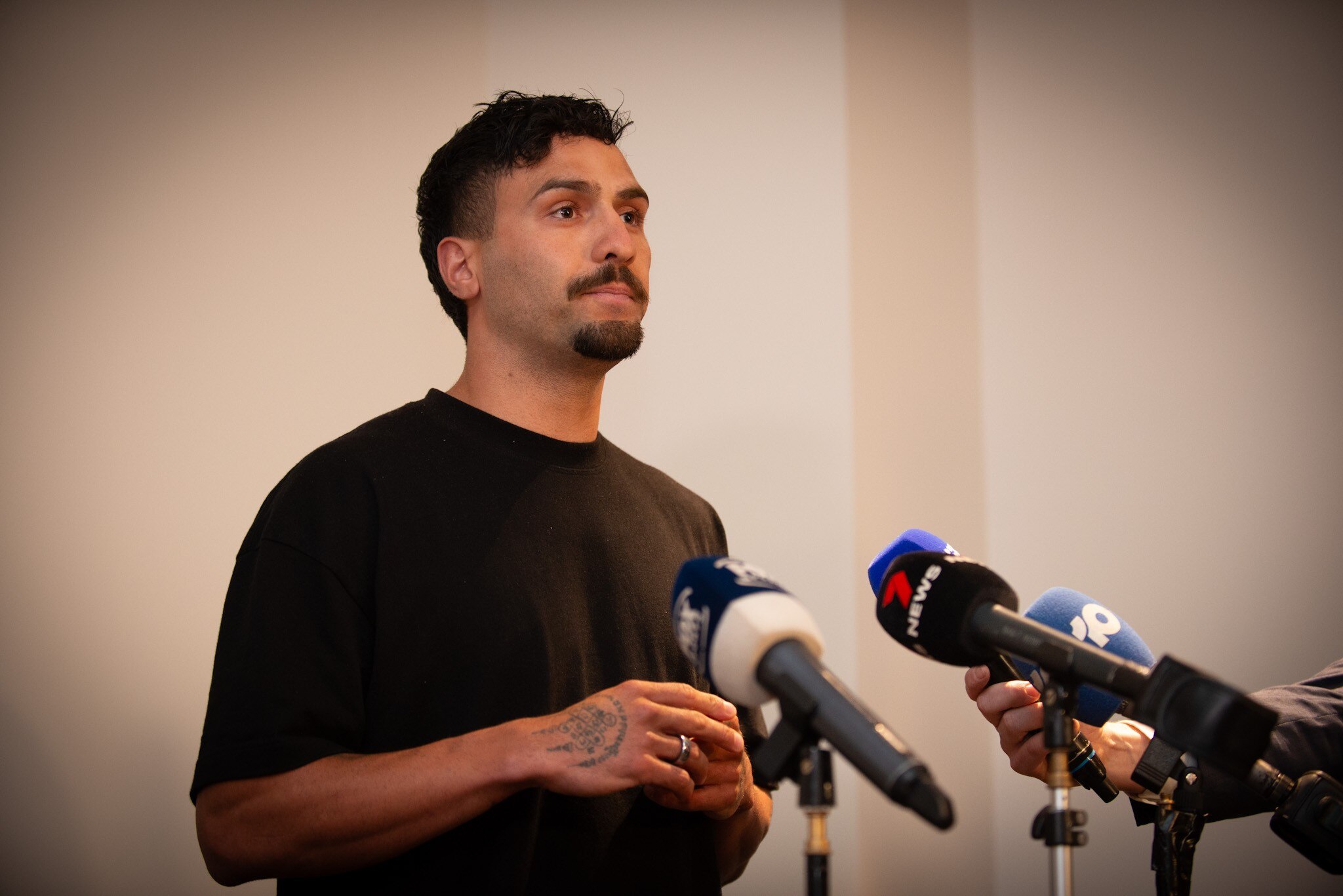 Serious Izak Rankine wears brown tee, stands in front of a host of microphones, wall behind is blank. He has a black goatee.