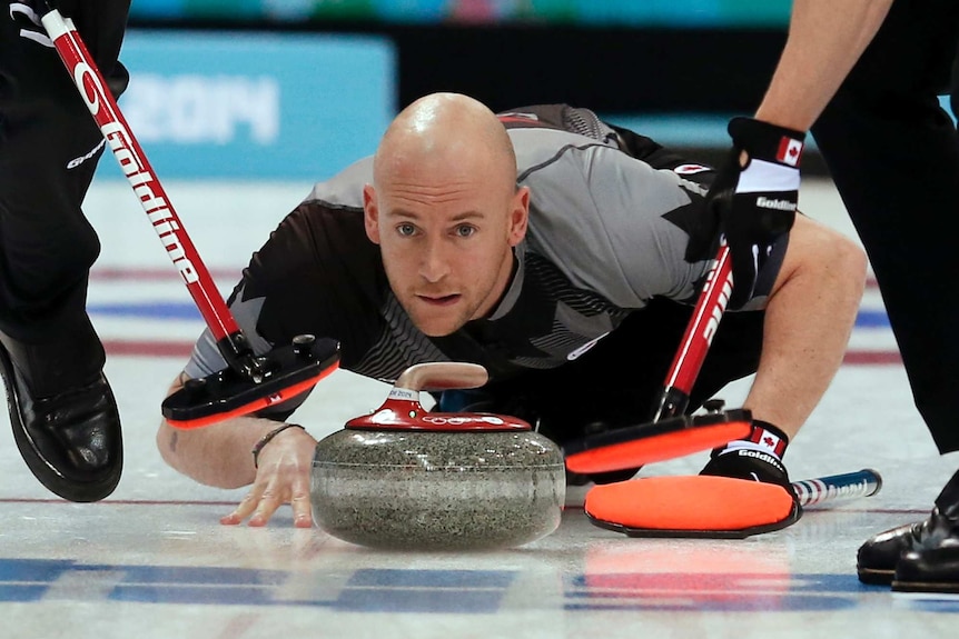A man in a grey and black sports jersey crouches on ice, with a curling stone and two brooms in front of his face.