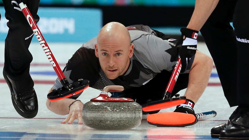 A man in a grey and black sports jersey crouches on ice, with a curling stone and two brooms in front of his face.
