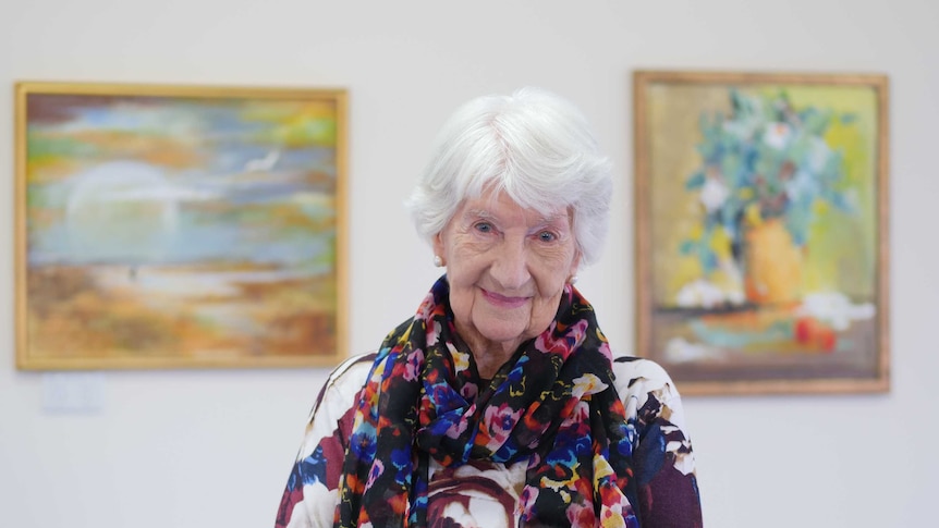 A senior woman in a colourful scarf and blouse smiling while two pieces of artwork hang blurred in the background.
