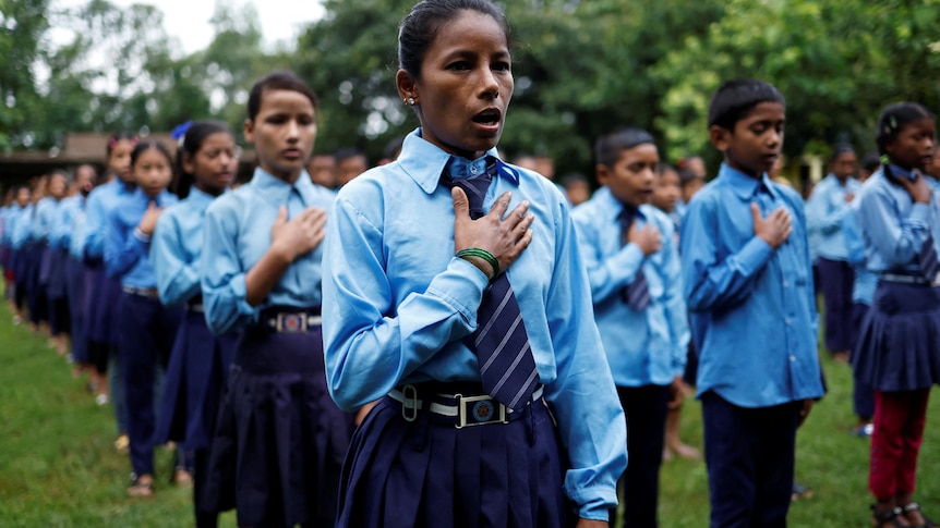 A woman stands in a line of school students all in uniform. All have one hand over chest and are singing