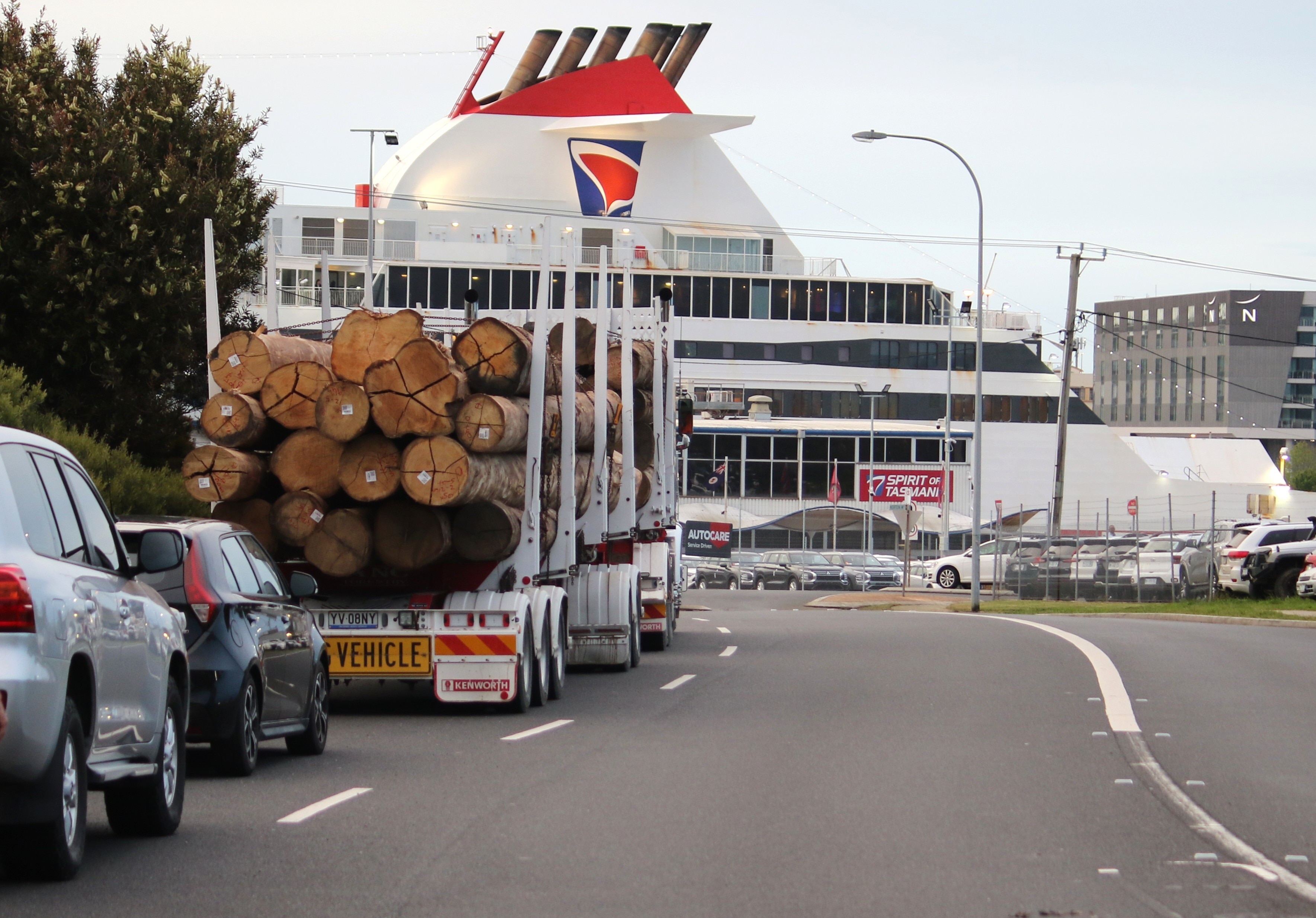 A semi-trailer loaded with logs in a traffic lane approaching a port. The large Spirit of Tasmania ship is in front of it.