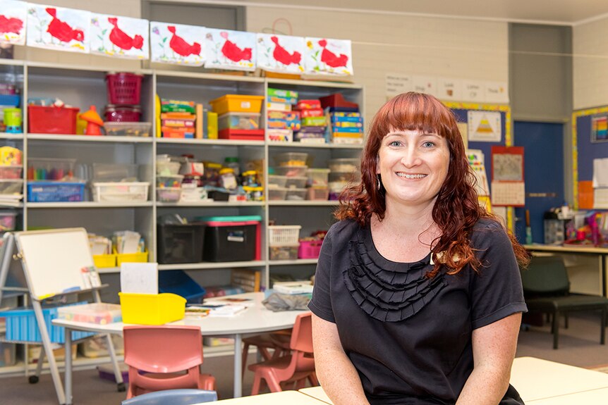 Woman with red hair sits with colourful, empty primary school classroom in background