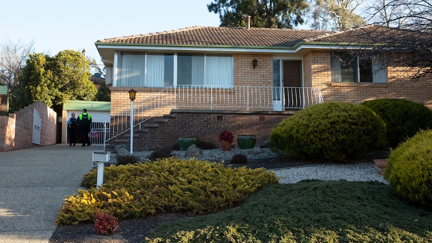 A brick house with steps up to a small porch. Police officers stand in the home's driveway.