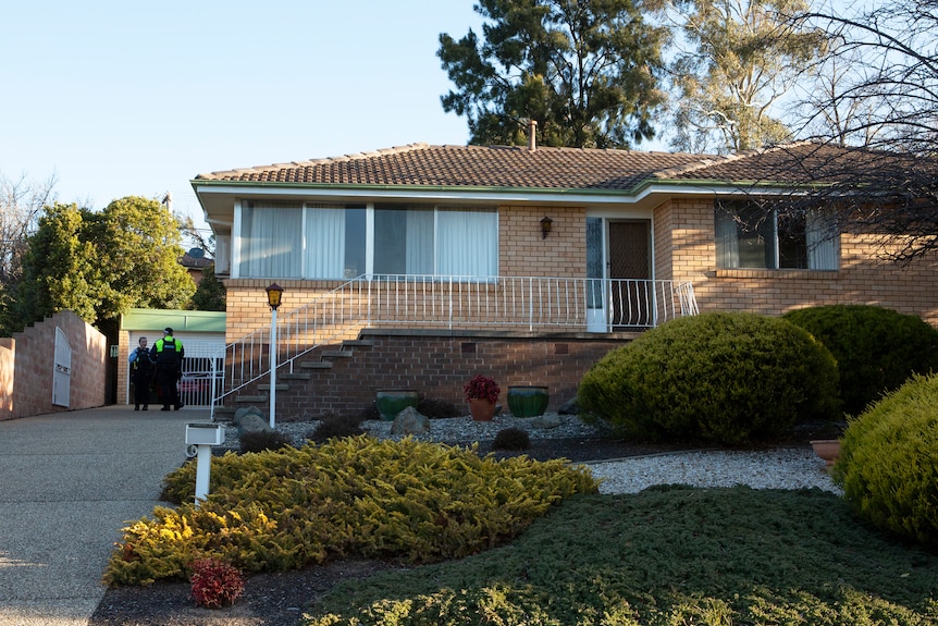 A brick house with steps up to a small porch. Police officers stand in the home's driveway.