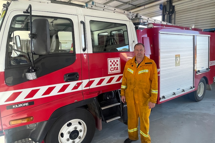 A man wearing yellow firefighting coveralls stands next to a red firetruck.