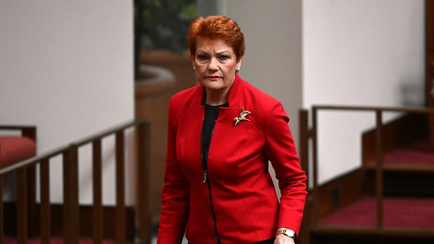 Pauline Hanson, dressed in a red jacket, wears a serious expression as she walks out of the Senate.