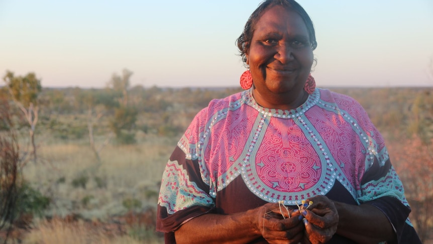 Indigenous woman wearing long pink earrings and brightly coloured kaftan smiling in front of bush landscape