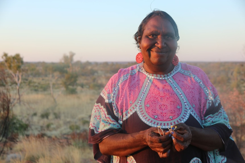 Indigenous woman wearing long pink earrings and brightly coloured kaftan smiling in front of bush landscape