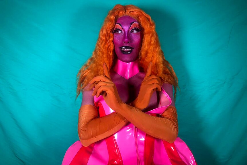 A drag queen in orange wig, plastic pink dress standing in front of a blue backdrop