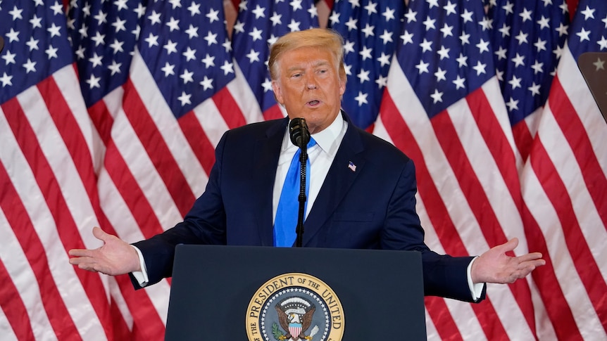 Donald Trump spreads his arms as he stands at the lecturn at the White House in front of American flags