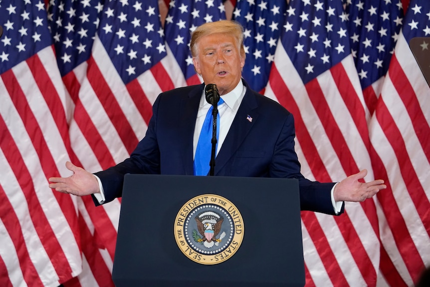 Donald Trump spreads his arms as he stands at the lecturn at the White House in front of American flags