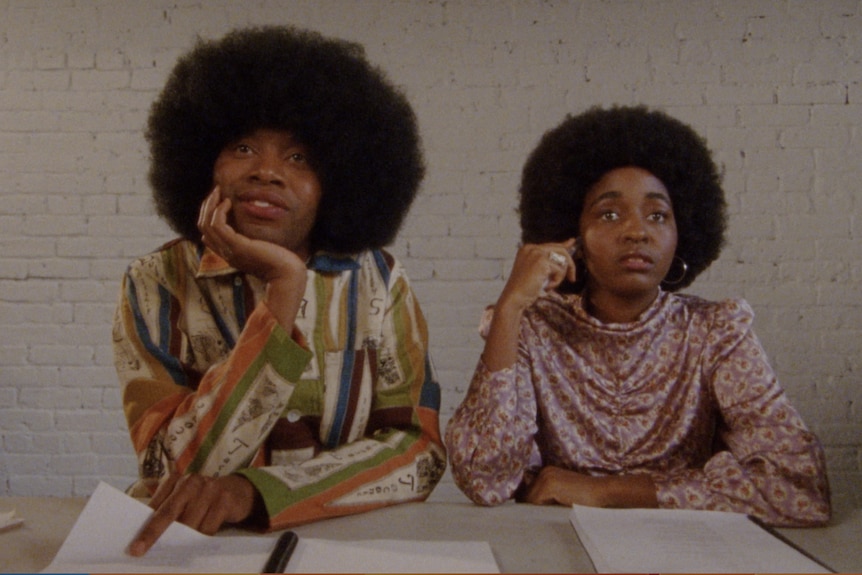 Two people with afros in bright clothing look forward from a casting table adorned with notes.