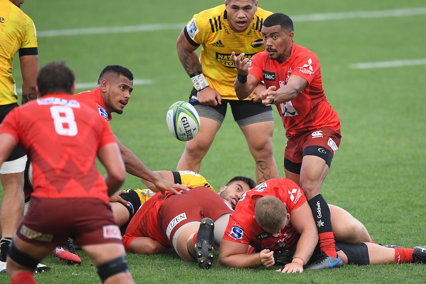 A Super Rugby player spins the ball towards a teammate, as other players lie on the ground during a match.