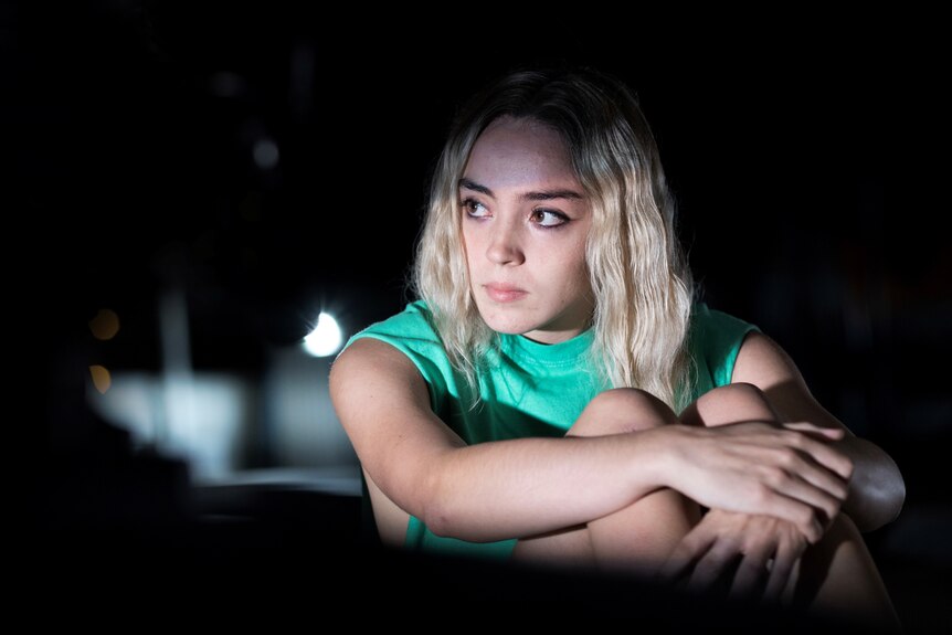 A young woman with blonde hair and green tank top sits in the dark but illuminated by light