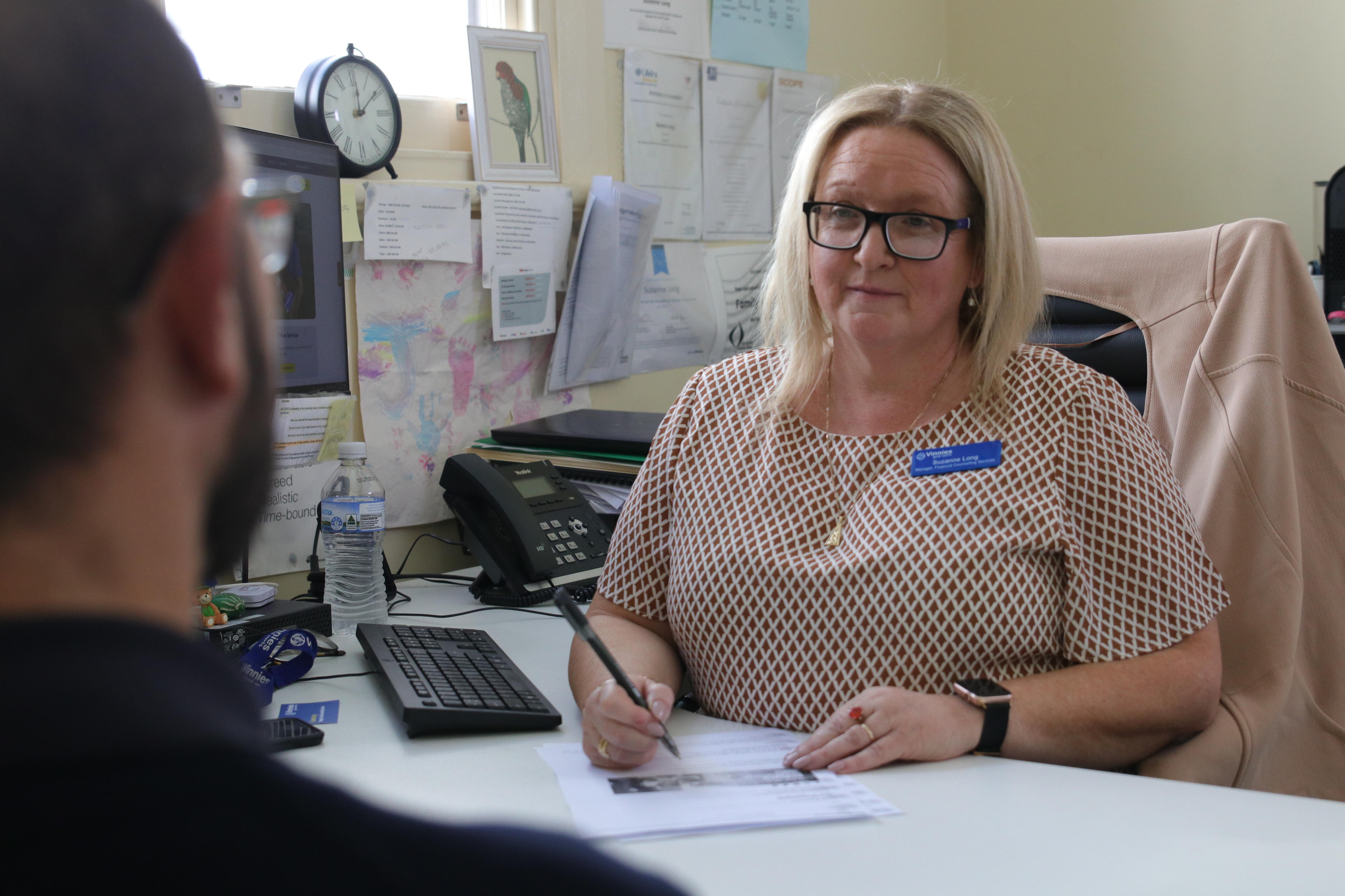 A middle-aged woman in an office, sitting at a desk, speaks to a man.