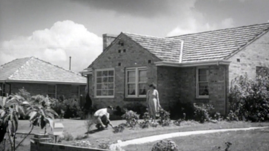 An old black and white photo of a man and woman pottering in the front garden of a suburban home