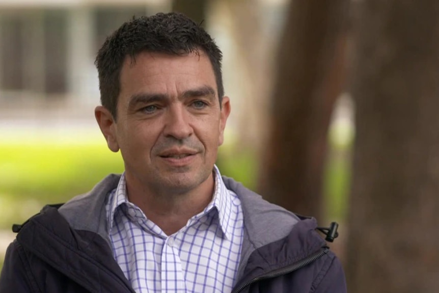 A dark-haired man in a business shirt and jacket smiles and looks at the camera.