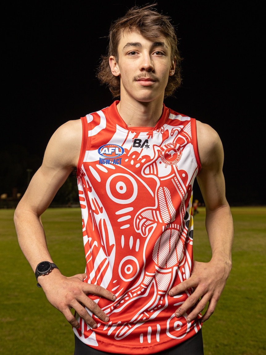 An Indigenous man with dark hair standing on a field at night with a red football guernsey on with his hands on his hips