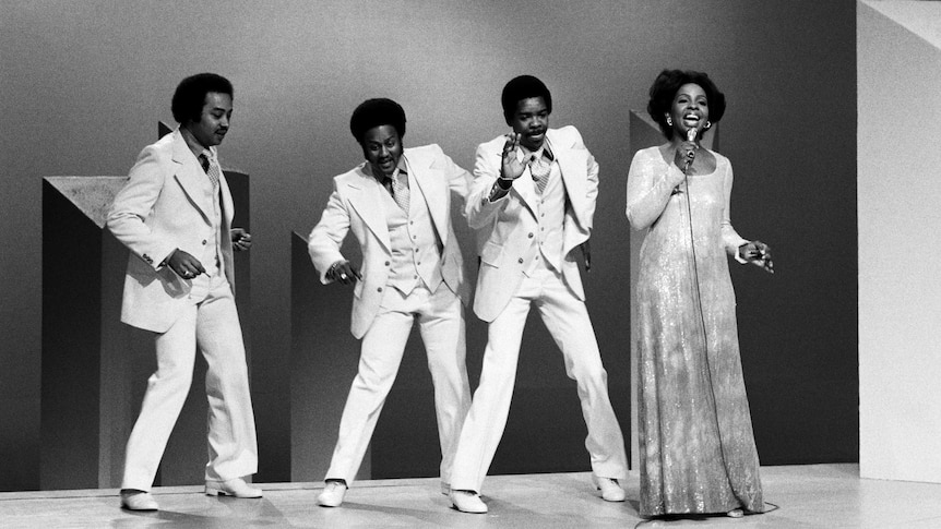 Gladys Knight and the Pips perform on stage. The men wear white suits and Gladys is holding a microphone wearing a long gown.