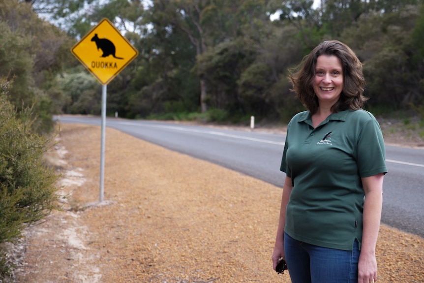 A woman standing near a road with a road sign warning drivers about quokkas