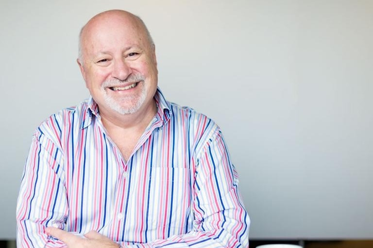 A man sits at a table a brightly lit room smiling at the camera with a blue mug in front of him.