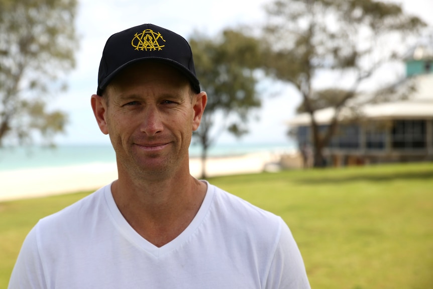 Headshot of a smiling man in a cap with greenery and a beach in the background.