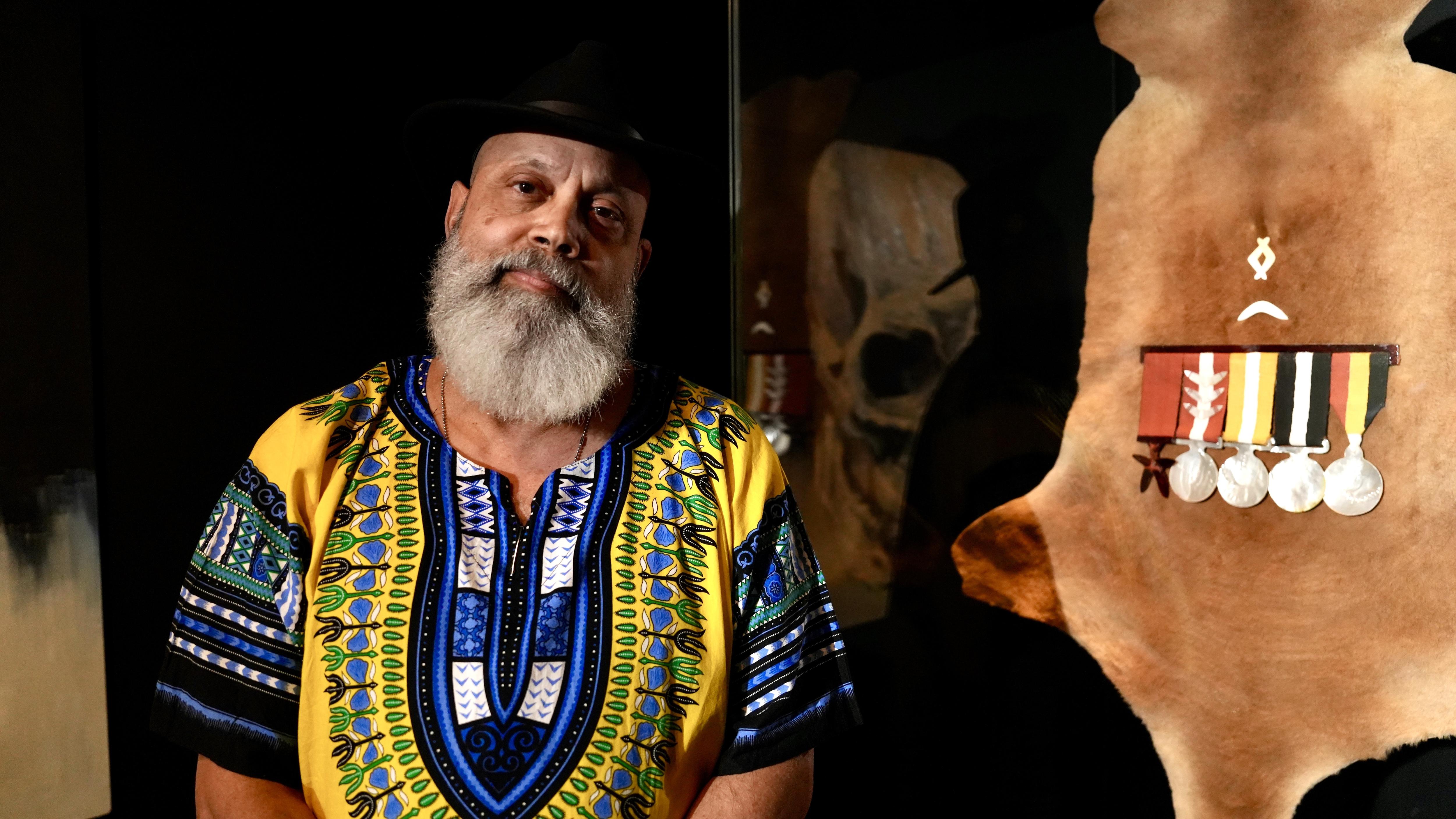 An Indigenous man stands in front of a kangaroo hide with First Nations-style artistic representation of Australian war medals.