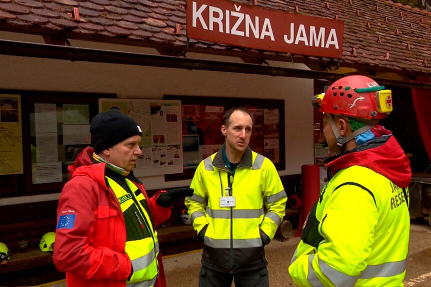 Man in yellow hi-vis and wearing red jackets gather in huddle outside of entry to Krizna Jama caves. 