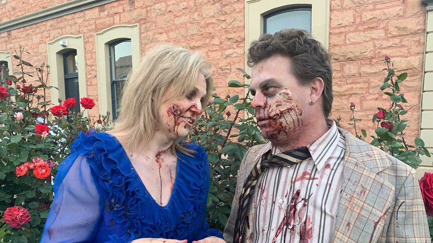 A blonde woman covered in fake blood and in a blue dress looks at a brown-haired man in a suit similarly covered in fake blood