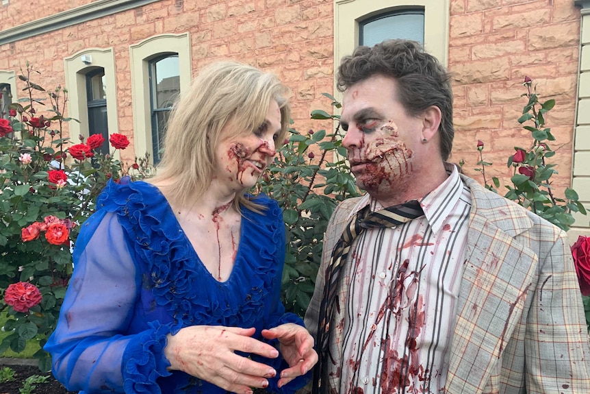 A blonde woman covered in fake blood and in a blue dress looks at a brown-haired man in a suit similarly covered in fake blood