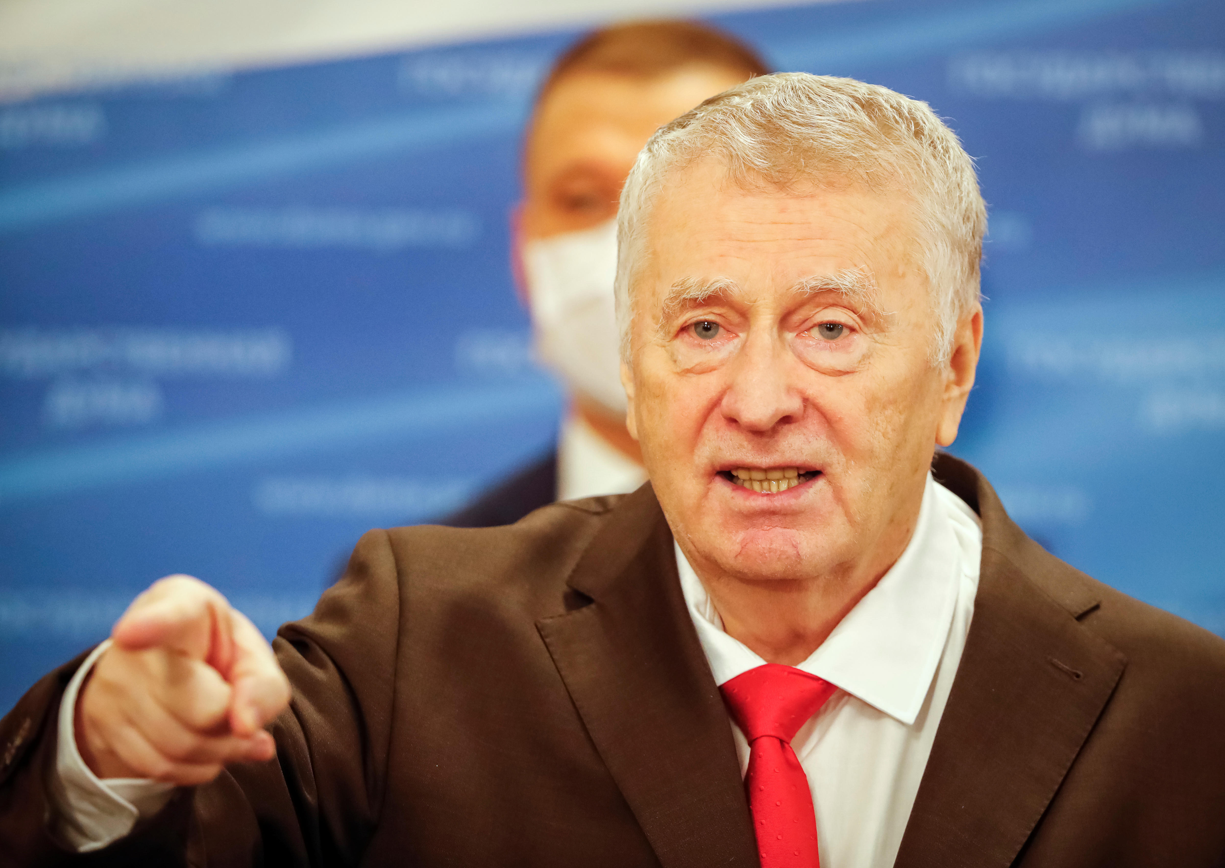 An old man in a suit with a red tie points as he speaks 