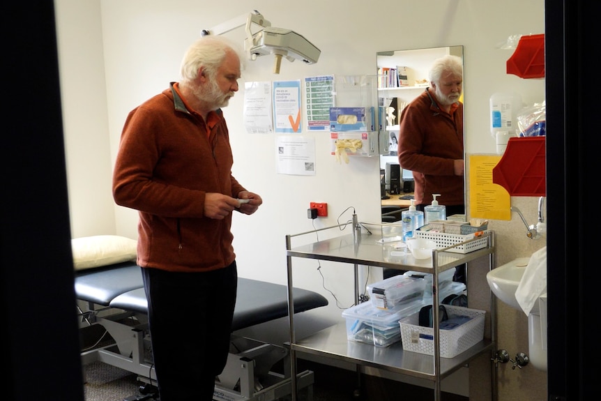 A man with gray hair and a gray beard wearing a red jumper stands in a GP consult room.