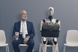 An older man in a suit sit next to a robot as they wait for a job interview