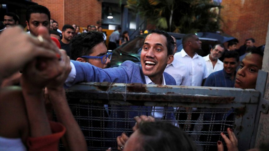 Smiling man with dark hair in blue suit jacket and white shirt reaches over a fence to touch hands of crowd