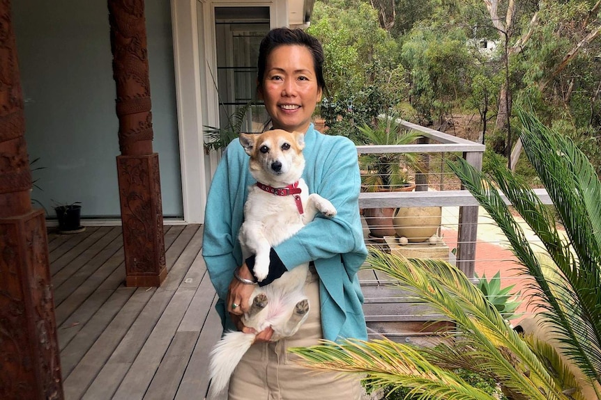 A portrait photo of a woman standing on a verandah holding her black and white dog and smiling.