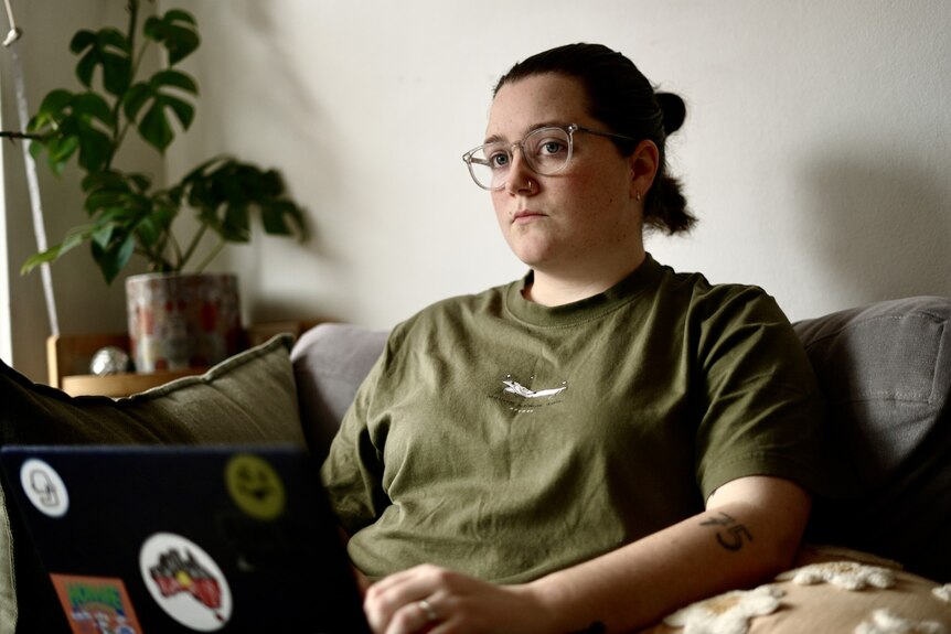 A young white woman with brown hair and glasses. She is sitting on a couch with a laptop