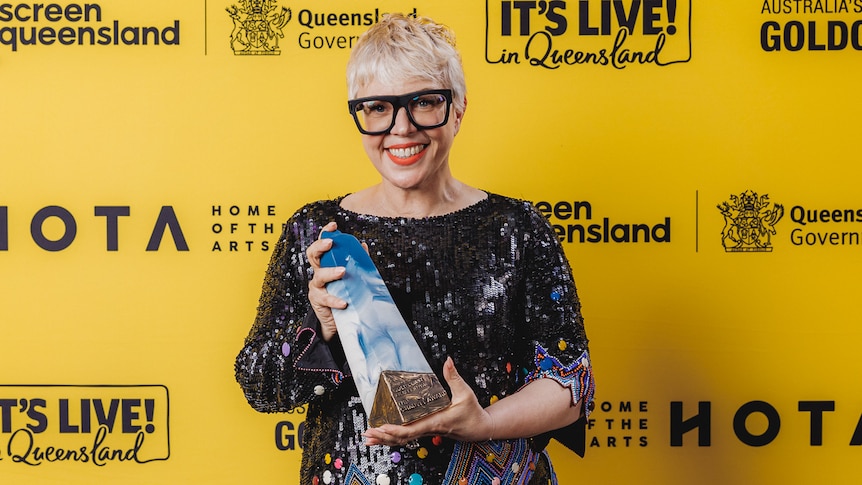 A woman with short blond hair and black rimmed glasses, wearing a black sequined dress holding an award.