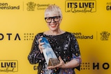 A woman with short blond hair and black rimmed glasses, wearing a black sequined dress holding an award.