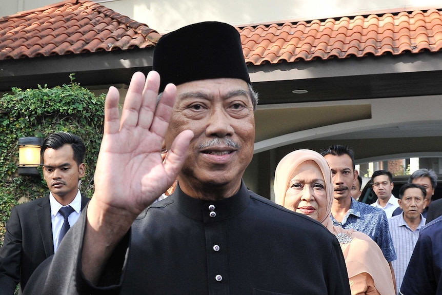 Muhyiddin Yassin waves and smiles as onlookers stand behind him at a house
