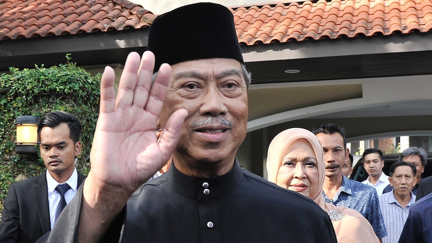 Muhyiddin Yassin waves and smiles as onlookers stand behind him at a house