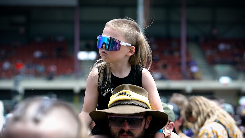 Boy with big sunglasses sitting on the shoulders of a man in the crowd. 