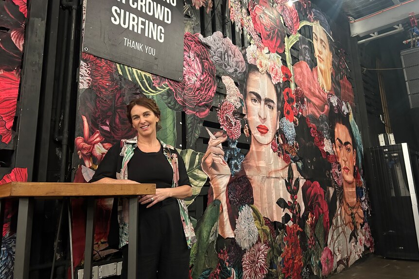 Una donna sta sorridente e si appoggia a un tavolo alto con un murale dipinto sul muro dietro di lei.