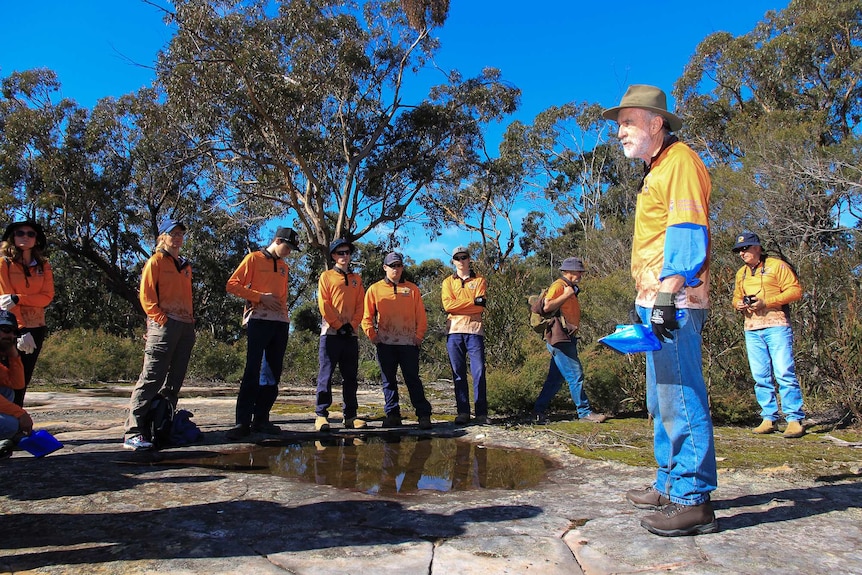 A man in an orange shirt with other people standing around in same shirts in a bush setting on a rock platform