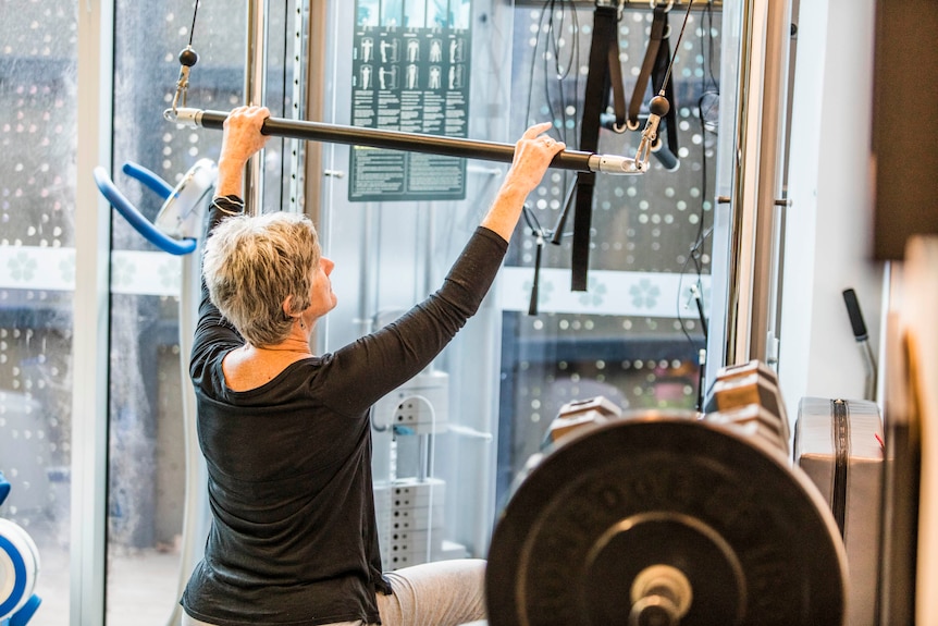 A woman in exercise clothes sits on a bench reaching up to a weights bar.