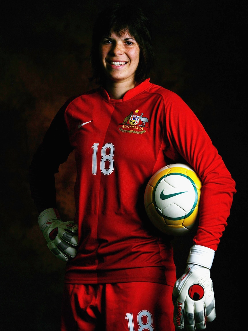 A woman athlete wearing red poses for a photo while holding a soccer ball under her arm