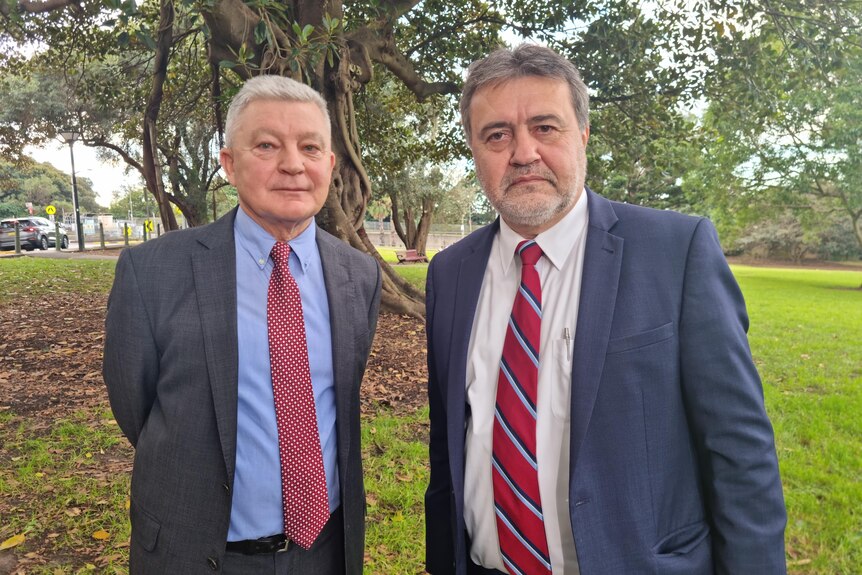 Two men wearing suits and ties stand side by side in a park, unsmiling