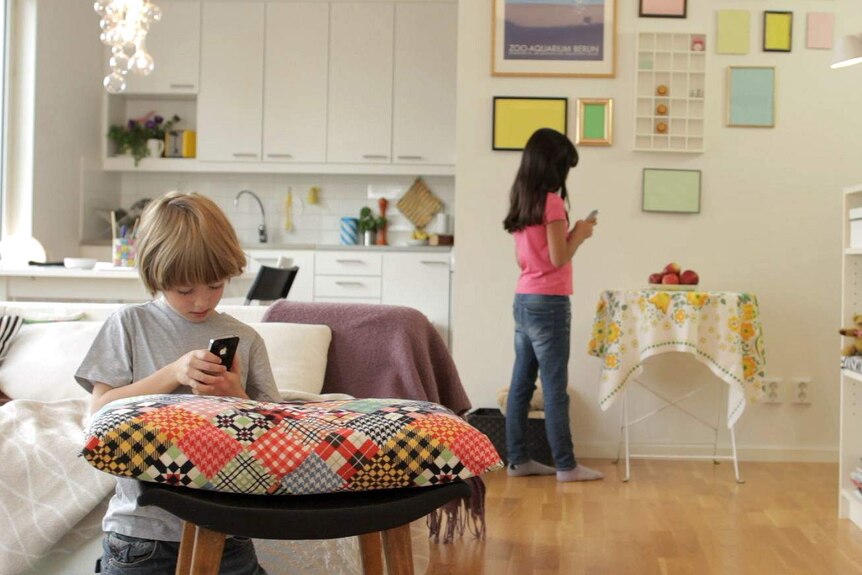 Two children stand inside their house using their mobile phones